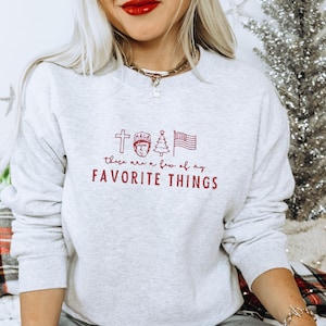 Puede incluir: Una sudadera gris claro con la frase "these are a few of my FAVORITE THINGS" en rojo. El diseño incluye una cruz, una cara con un sombrero "MAGA", un árbol de Navidad y una bandera estadounidense.