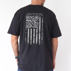 May include: Black t-shirt with a distressed American flag design on the back. The flag is composed of vertical stripes and a QR code in place of the stars. The text "THANK YOU FOR YOUR ATTENTION TO THIS MATTER" is above the flag.