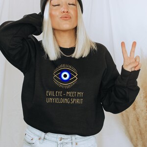 May include: Black sweatshirt with a gold evil eye graphic and the text "Evil Eye - Meet My Unyielding Spirit".