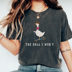 Op de afbeelding: Een donkergrijs T-shirt met een cartoonillustratie van een gans die een cowboyhoed en een bandana draagt en een step rijdt. De tekst "THE HELL I WON'T" is onder de afbeelding gedrukt.