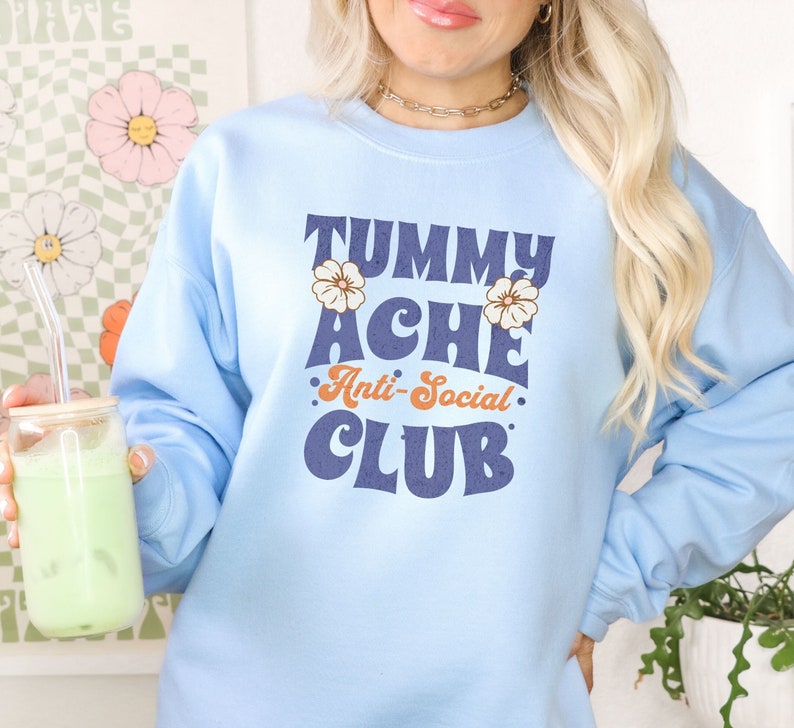 May include: A light blue sweatshirt with the text "Tummy Ache Anti-Social Club" in a retro font. The text is in navy blue with orange accents. There are two small white flowers on either side of the text.