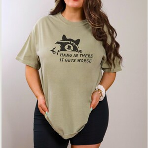 Peut inclure: T-shirt vert olive avec un dessin de raton laveur noir et le texte "Hang in there, it gets worse". Le t-shirt a des manches courtes et une coupe ample. Le motif est imprim&eacute; sur le devant.