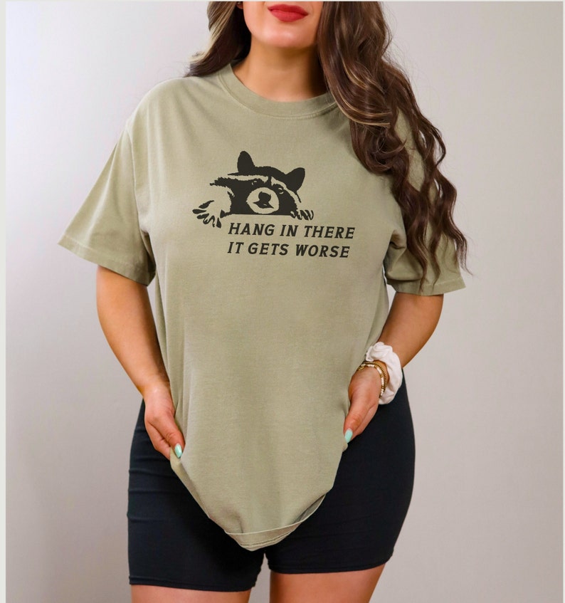 Peut inclure: T-shirt vert olive avec un dessin de raton laveur noir et le texte "Hang in there, it gets worse". Le t-shirt a des manches courtes et une coupe ample. Le motif est imprim&eacute; sur le devant.