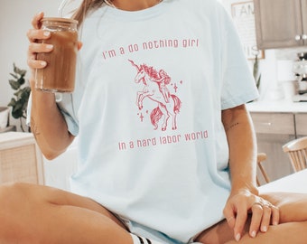 May include: A light blue t-shirt with a red graphic of a unicorn with a woman riding it. The text "I'm a do nothing girl in a hard labor world" is printed on the shirt.