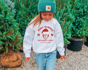 May include: A white sweatshirt with red text that says "Merry Trumpmas and a Happy 4 Years Let Freedom Ring". A cartoon image of Donald Trump wearing a Santa hat is in the center of the text. The sweatshirt is being worn by a child wearing a green beanie and blue jeans.