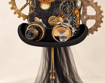 Peut inclure: Un haut-de-forme noir avec une bande en tissu à motifs célestes, orné de grands engrenages dorés et noirs, de lunettes en laiton avec un seul verre et d'une paire de petites lunettes circulaires. Un voile en tulle noir pend du bord.