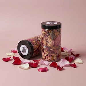 May include: Two clear cylindrical containers filled with dried rose petals, one upright and one on its side. The containers have black lids with a circular label. Scattered rose petals in shades of red, pink, and white surround the containers on a pink surface.