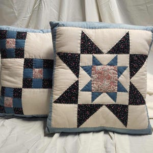 Puede incluir: Dos cojines cuadrados con diseños de patchwork. Uno muestra un patrón de estrella con tela color crema, azul y floral negro. El otro cojín presenta una cuadrícula de cuadrados en patrones a juego de color crema, azul y floral negro.