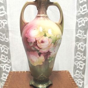 Può includere: Un vaso in porcellana a due manici caratterizzato da un motivo floreale dipinto a mano con rose rosa e gialle su uno sfondo sfumato che passa da un crema tenue nella parte superiore a un verde oliva scuro alla base, con accenti dorati.