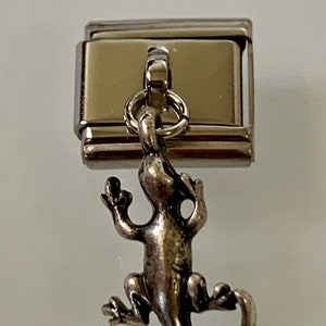 Puede incluir: Un charm plateado con un colgante detallado de lagarto suspendido de una base metálica rectangular. El lagarto tiene una cola curvada y está unido a la base con un pequeño anillo. El charm está diseñado para una pulsera o collar.