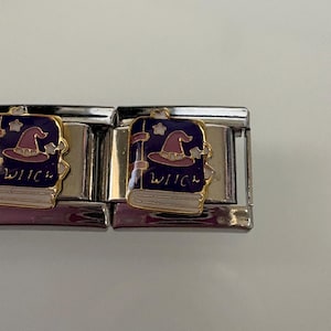 Könnte beinhalten: Zwei silberfarbene Charm-Links mit Hexen-Motiv. Jeder Charm zeigt ein lila Buch mit Hexenhut und Sternen sowie das Wort "WITCH" in Weiß. Die Charms sind vor einem hellen Hintergrund platziert.