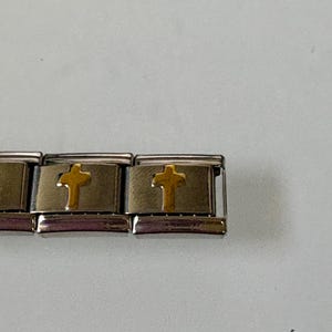 Op de afbeelding: Een zilverkleurige armband met goudkleurige kruisbedels. De kruisen zijn gecentreerd op rechthoekige schakels. De armband ligt op een lichtgrijze achtergrond.