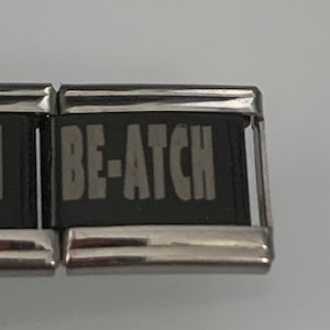 Może przedstawiać: Srebrna bransoletka z czarującą zawieszką, na której widnieje napis "BE-ATCH" białymi literami.