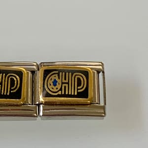Op de afbeelding: Twee zilveren en gouden armbandbedels met zwart emaille en gouden accenten. De bedel rechts zegt "CHIP" met een blauwe ster in het midden.
