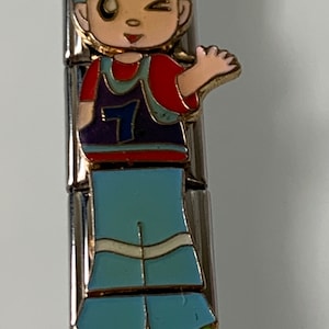 May include: A charm bracelet charm featuring a cartoon boy wearing a blue shirt, red and purple vest with the number 7, blue pants, and green shoes. The boy is winking and has a smile on his face.