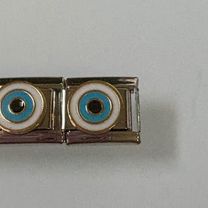 Könnte beinhalten: Nahaufnahme von zwei silberfarbenen Charm-Gliedern, jedes mit einem weiß-gold umrandeten Augendesign mit türkisfarbenem Zentrum. Die Charms sind verbunden und zeigen eine Schmuckkomponente.