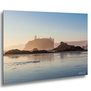 Op de afbeelding: Een landschapsfoto van een strand bij zonsondergang. De afbeelding toont een kalme zee die de kleuren van de lucht weerspiegelt, met grote rotsen en een klein eiland in de verte. De lucht gaat over van blauw naar oranje.