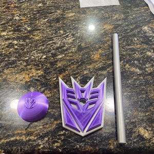 May include: A purple and silver Decepticon logo, a purple disc with a Decepticon logo, and a silver rod are displayed on a dark countertop. The Decepticon logo is a stylized emblem with a purple center and silver outline.