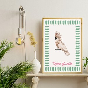Op de afbeelding: Een ingelijste print met een aquarel illustratie van een roze en witte kaketoe. Het kunstwerk heeft een groene penseelstreek rand en de tekst "Queen of noise" in roze.
