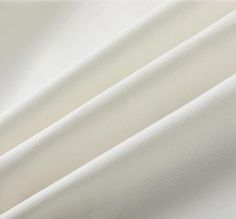 K&ouml;nnte beinhalten: Nahaufnahme eines cremefarbenen Stoffes mit diagonalen Falten. Der Stoff hat eine subtile Textur und scheint ein mittelschweres Material zu sein. Die Falten erzeugen Schatten und Lichtreflexe und verleihen dem Bild Tiefe. Die Farbe des Stoffes ist ein weiches, cremiges Wei&szlig;.