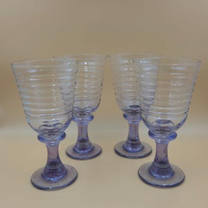 May include: A set of four clear glass goblets featuring horizontal ribbed detailing along the upper bowls. Each glass has a thick, sturdy stem and a circular base, both of which are tinted with a subtle, light purple hue.