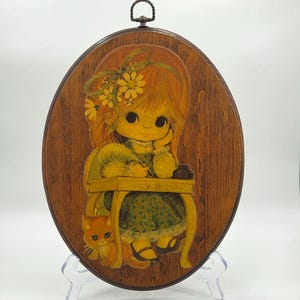 May include: Oval wooden wall hanging featuring a cartoon girl with orange hair, a green dress, and a yellow desk. A small orange cat sits beside her. The artwork has a vintage, retro style with a metal hanging loop at the top.