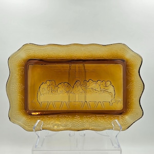May include: Amber-colored glass serving tray with a rectangular center featuring an etched depiction of The Last Supper. The tray has a decorative, scalloped edge with a textured pattern. The tray is supported by a clear plastic stand.