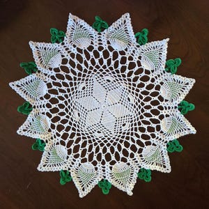 May include: A round white crochet doily featuring a central star pattern, intricate openwork lace, and a scalloped border. The edge is decorated with alternating white and light green triangular motifs and small, bright green three-leaf clover shapes.