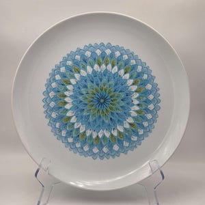 May include: White ceramic plate with a central floral design in shades of blue, green, and white. The plate has a wide rim and is displayed on a clear stand. The floral pattern is symmetrical and detailed.