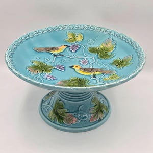 May include: A turquoise ceramic pedestal cake stand with a detailed design. The stand features two yellow and brown birds, green leaves, and purple grapes. The rim has a decorative border. The base is also decorated with leaves and vines.