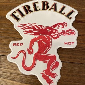 Op de afbeelding: Witte patch met het woord "FIREBALL" in zwarte en gouden letters. Een rode, gespierde, demonachtige figuur met vlammen rent over de patch. De woorden "RED" en "HOT" staan aan weerszijden van de figuur.