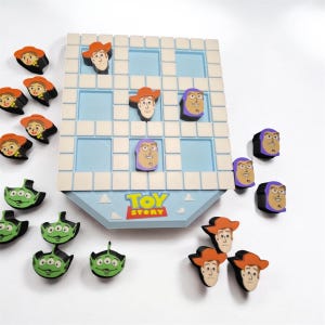 May include: A Toy Story themed tic-tac-toe game board with a light blue grid and white borders. The set includes several character game pieces featuring Woody, Buzz Lightyear, and green aliens. The board displays the Toy Story logo in red and yellow.