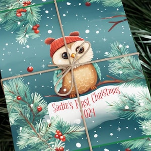 May include: A blue and white Christmas wrapping paper with a cute cartoon owl wearing a red hat. The paper has a snowy background with pine branches and red berries. The text on the paper reads "Sadie's First Christmas 2024".