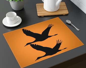 May include: Orange placemat with two black silhouettes of cranes flying in the air.