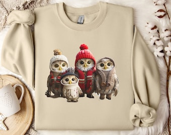 May include: A beige sweatshirt with a graphic print of four owls wearing winter hats and scarves. The owls are arranged in a row, with the largest owl in the center.