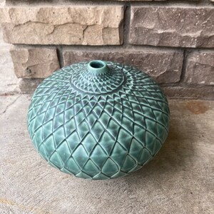 Pode incluir: Um vaso de cerâmica verde-azulado com um padrão geométrico texturizado. O vaso tem uma forma arredondada com uma pequena abertura no topo. A superfície é coberta por um design de diamante repetido, criando uma textura tátil e visualmente atraente.