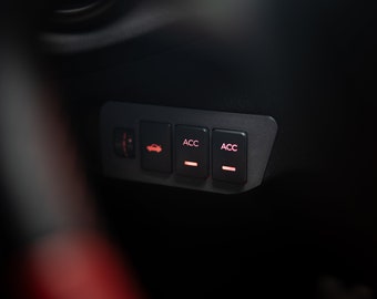 May include: A black panel featuring illuminated red buttons, possibly for a car's interior. The panel has buttons marked "ACC" and a car symbol, indicating control over vehicle functions. The lighting is low, creating a focused view.