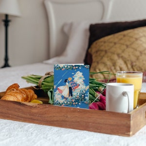May include: A wooden tray with a white mug, a glass of orange juice, a croissant, a bouquet of tulips, and a card with a blue and white illustration of a couple dancing.