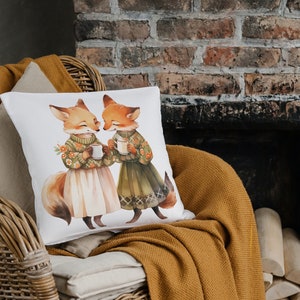 May include: A white throw pillow with a watercolor illustration of two foxes holding mugs. The foxes are wearing sweaters and skirts. The background is a brick wall.