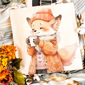 May include: A tote bag featuring a cartoon fox wearing a knitted sweater and a hat, holding a cup of coffee. The fox has a fluffy tail and is surrounded by autumnal colors.