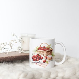 May include: White ceramic mug featuring a watercolor illustration of cherries in a basket and on a checkered cloth. A small cup and a basket filled with red cherries are also depicted. A candle and decorative sprig are in the background.