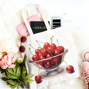 May include: A white tote bag featuring a watercolor illustration of a bowl overflowing with ripe red cherries. The bag contains a pink notebook, a black notebook with gold lettering, and a smartphone. Pink sunglasses, flowers, and shoes are arranged around the bag.