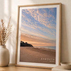 May include: A framed print of Exmouth beach in Devon, showing a sunset sky with wispy clouds. The beach is sandy, with a small hill on the left. The print is in a light wood frame, with the text "EXMOUTH DEVON" at the bottom.