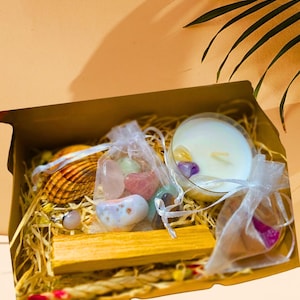 May include: A gift box filled with a variety of items. Includes a seashell, a ring with a pink stone, a small ceramic bowl, a wooden stick, a candle, and small bags of colorful crystals. The box is lined with straw.