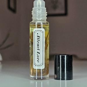 May include: A clear glass bottle with a black roll-on top filled with a light yellow liquid. The bottle has a white label with the text "Attract Love" in black.