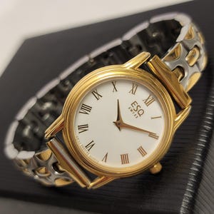 May include: A gold and silver two-tone watch with a white face and Roman numeral hour markers. The watch has a gold band with a clasp.