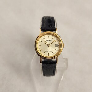 K&ouml;nnte beinhalten: Eine goldfarbene Lorus-Armbanduhr mit schwarzem Lederarmband. Das Zifferblatt der Uhr ist goldfarben mit schwarzen Zeigern und einem Datumsfenster. Die Uhr ist wasserdicht und hat ein klassisches Design.