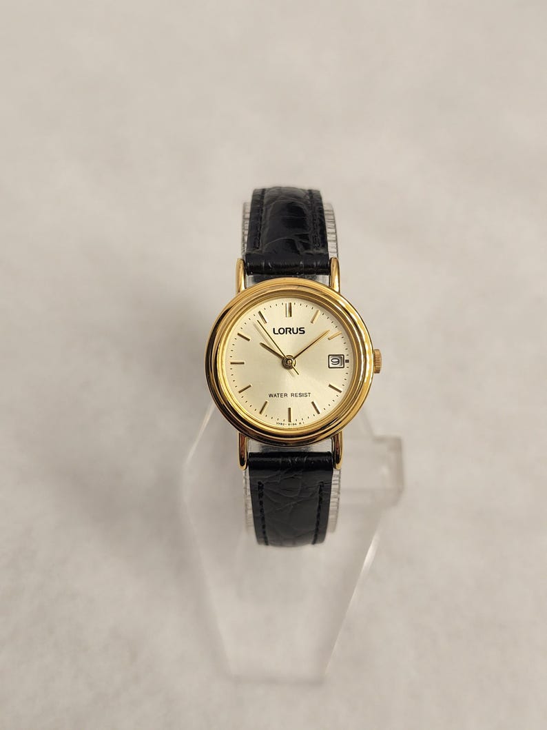 K&ouml;nnte beinhalten: Eine goldfarbene Lorus-Armbanduhr mit schwarzem Lederarmband. Das Zifferblatt der Uhr ist goldfarben mit schwarzen Zeigern und einem Datumsfenster. Die Uhr ist wasserdicht und hat ein klassisches Design.