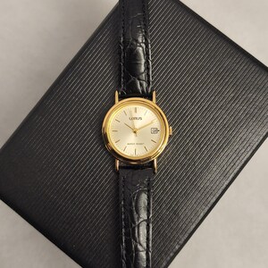 K&ouml;nnte beinhalten: Eine goldfarbene Lorus-Uhr mit einem schwarzen Lederarmband. Das Zifferblatt der Uhr ist hell mit goldenen Zeigern und Markierungen. Das Armband hat ein strukturiertes, krokodilartiges Muster. Die Uhr ist wasserdicht.