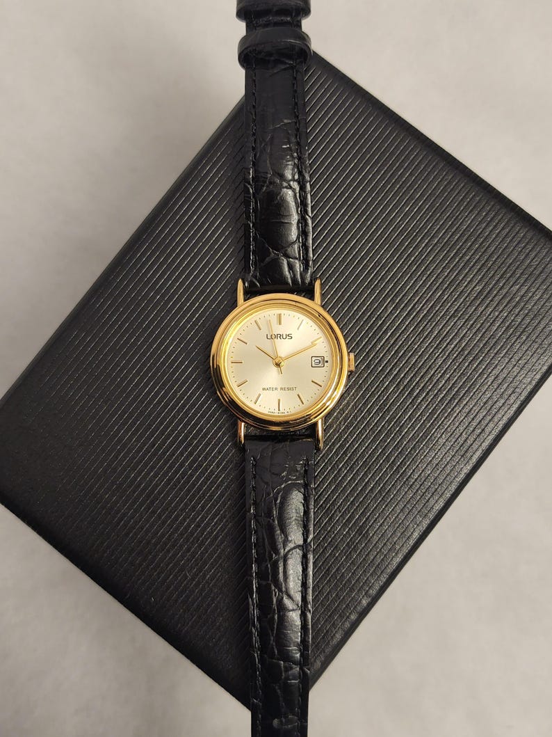K&ouml;nnte beinhalten: Eine goldfarbene Lorus-Uhr mit einem schwarzen Lederarmband. Das Zifferblatt der Uhr ist hell mit goldenen Zeigern und Markierungen. Das Armband hat ein strukturiertes, krokodilartiges Muster. Die Uhr ist wasserdicht.